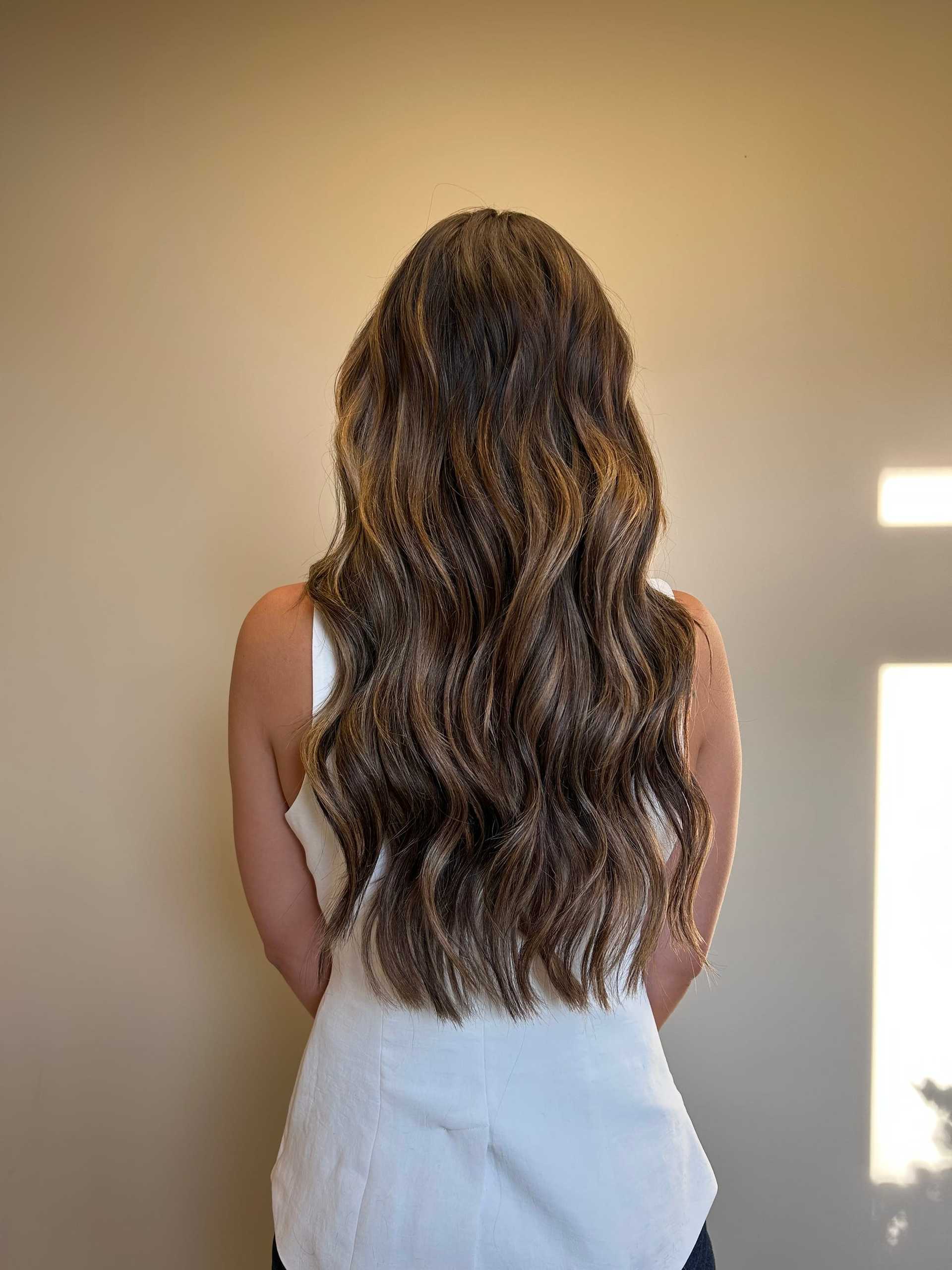 Long wavy brown hair cascading down woman's back against a plain beige background.